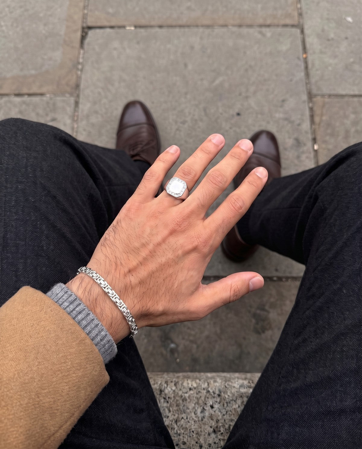 A man's hand over charcoal trousers and a camel coat, wearing the King's Byzantine silver bracelet and the Frontier Signet ring